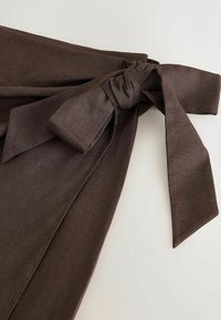Pantalon en tissu marron foncé avec une large ceinture nouée en noeud papillon sur un fond clair.