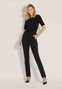 Zwarte, strakke broek met studded accenten aan de zijkanten, gecombineerd met een zwart topje met korte mouwen. Model staat in hoge hakken tegen een neutrale achtergrond.
