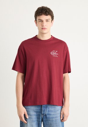 T-shirt in cotone bordeaux con maniche corte, scollatura rotonda e un logo bianco a contrasto sul lato sinistro del petto. Vestibilità casual e texture morbida.