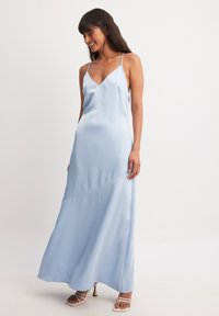 Šviesiai mėlyna satino maksi suknelė su V formos iškirpte ir plonomis petnešėlėmis, turinti laisvą siluetą ir subtilias įstrižaines siūles.