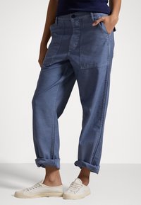 Pantalon en denim bleu avec une coupe décontractée, doté de plusieurs poches, de poignets roulés et d'une texture subtile. Associé à des baskets de couleur claire.