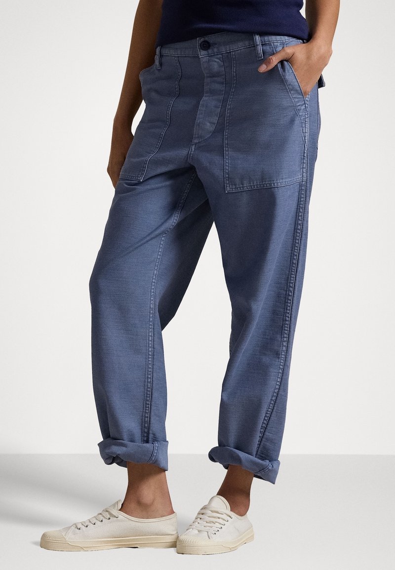 Pantalon en denim bleu avec une coupe décontractée, doté de plusieurs poches, de poignets roulés et d'une texture subtile. Associé à des baskets de couleur claire.
