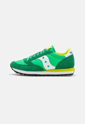 Chaussure de running verte et jaune avec logo blanc sur le côté, lacets verts, semelle à crampons noire, et languette blanche au talon sur fond uni.
