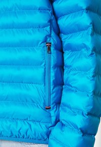 Giacca a vento blu con design a palchettone orizzontale. Presenta una tasca con zip, una texture liscia e un dettaglio del logo sull'orlo.