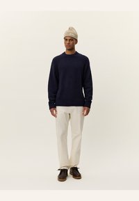 Navy blue knitted sweater with ribbed cuffs and hem, paired with cream-colored trousers and brown lace-up shoes. Beige beanie worn on head.