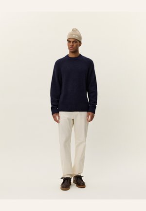 Navy blue knitted sweater with ribbed cuffs and hem, paired with cream-colored trousers and brown lace-up shoes. Beige beanie worn on head.
