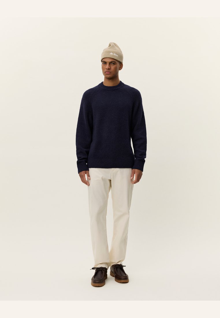 Navy blue knitted sweater with ribbed cuffs and hem, paired with cream-colored trousers and brown lace-up shoes. Beige beanie worn on head.