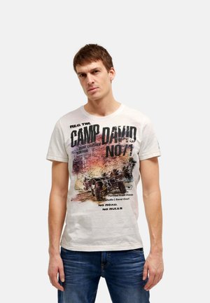 Young man with short brown hair wearing a white graphic t-shirt featuring a dune buggy and text, paired with blue jeans, standing against white background.