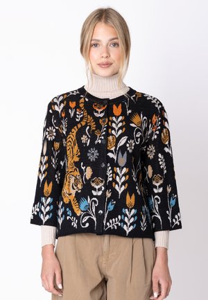 Donna che indossa un cardigan nero con bottoni con motivi floreali e di tigre colorati sopra un maglione beige a collo alto e pantaloni beige.