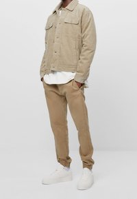 Béžová manšestrová bunda s límečkem z beránčí kožešiny, spárovaná s béžovými zúženými kalhotami a bílými teniskami. Oblečení doplňuje vrstvený bílý top pod bundou.