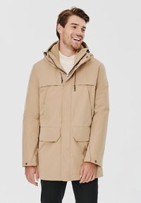 Beżowa wodoodporna parka z kapturem, z zamkiem błyskawicznym z przodu, dwoma dużymi kieszeniami z przodu, regulowanymi sznurkami i gładką teksturą.