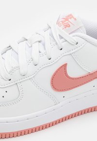 Bílé tenisky Nike Air Force 1 s růžovým logem swoosh, odpovídající růžovou podešví a bílými tkaničkami na čistém bílém pozadí.