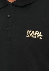 Juodas polo marškinėliai su trimis sagomis ir auksiniu KARL LAGERFELD logotipu kairėje krūtinės pusėje, kuriame yra mažas silueto detalė.