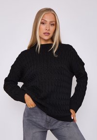 Schwarzer gestrickter Pullover mit strukturiertem Muster, Rundhalsausschnitt und gerippten Bündchen. Kombiniert mit grauen Jeans, die eine lockere Passform haben.