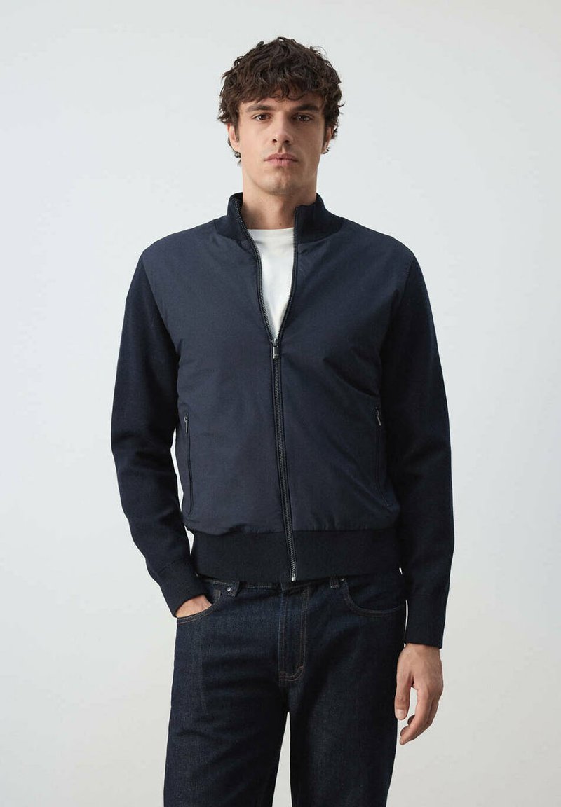 Jeune homme aux cheveux bouclés portant une veste zippée bleu marine foncé sur une chemise blanche et un jean bleu foncé, debout devant un fond uni.