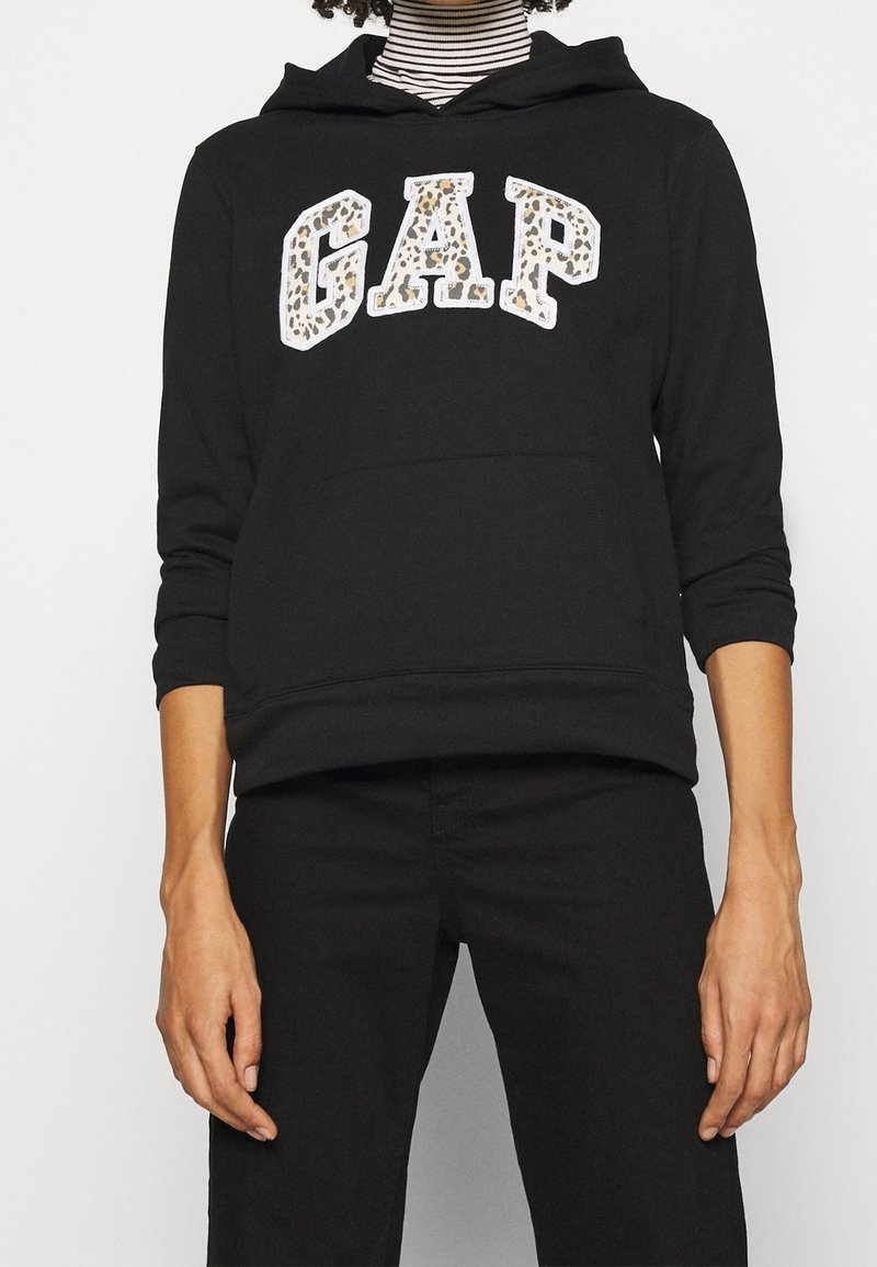 Personne portant un sweat à capuche noir avec imprimé léopard "GAP" logo, pantalon noir et un col roulé rayé noir et blanc en dessous.