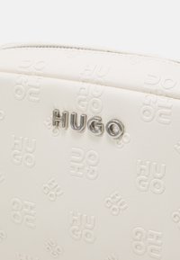 Hvid tekstureret lædertaske med præget "HUGO" logo. Sølvfarvet hardware og lynlåslukning øverst.