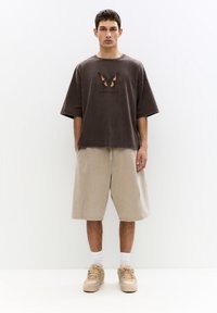 T-shirt oversize grigia scura con grafica di una farfalla; abbinato a pantaloni corti beige e sneakers beige, completato da calze bianche.
