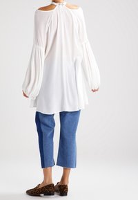 Vit långärmad blus med öppen axeldesign och voluminösa ärmar, kombinerad med blå cropped jeans och bruna skor med leopardmönster.