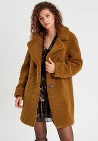 Manteau en peluche brun avec un col, design surdimensionné, tissu texturé, fermeture à bouton unique, porté sur une robe fleurie.