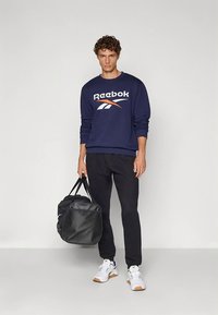 Marineblauwe sweatshirt met wit Reebok-logo, gecombineerd met zwarte broek en witte sportschoenen. Het model houdt een zwarte reistas vast.