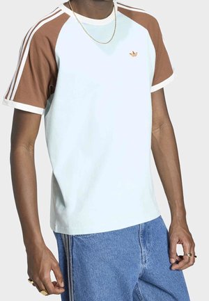Personne portant un t-shirt à manches courtes Adidas blanc et marron et un jean bleu, avec une chaîne en or et des bagues à la main gauche.
