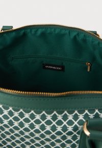 Πράσινη τσάντα χειρός με γεωμετρικό λευκό σχέδιο, κλείσιμο με φερμουάρ και επενδεδυμένο εσωτερικό που διαθέτει μικρή τσέπη με φερμουάρ και ετικέτα μάρκας.