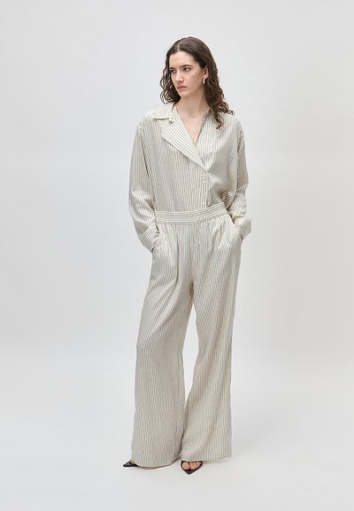 Γυναίκα με μπούκλες, φορώντας ένα κρεμ πουα πουκάμισο με κοντό μανίκι και φαρδιά παντελόνια, στέκεται με τα χέρια στις τσέπες σε απλό φόντο.