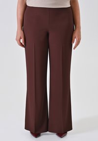 Persona che indossa pantaloni a vita alta e larghi di colore marrone scuro abbinati a scarpe a punta rosso scuro, in piedi davanti a uno sfondo chiaro e uniforme.