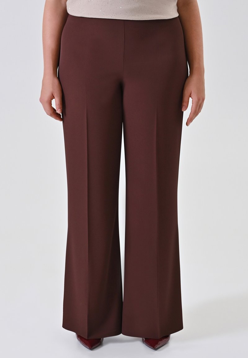 Persona che indossa pantaloni a vita alta e larghi di colore marrone scuro abbinati a scarpe a punta rosso scuro, in piedi davanti a uno sfondo chiaro e uniforme.
