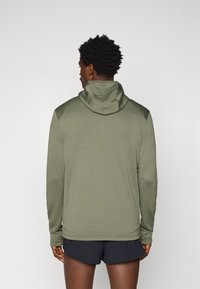 Maglione a maniche lunghe con cappuccio di colore verde oliva, realizzato in tessuto testurizzato, design aderente e maniche raglan. Pantaloni corti neri visibili sotto l'orlo.