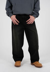 Pantaloni in denim nero con vestibilità comoda, indossati con una felpa con cappuccio grigio scuro e scarpe marrone chiaro dal design stile mocassino.