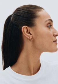 Goldfarbene Creolen in verschiedenen Größen, mit einem eleganten Design und poliertem Finish sowie einer glatten Textur, die an dem Ohr eines Modells getragen werden.
