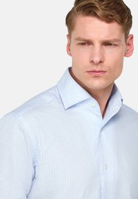 Chemise en coton bleu clair à rayures, avec un col boutonné. Le tissu semble lisse, présentant de subtiles rayures verticales et un design ajusté.