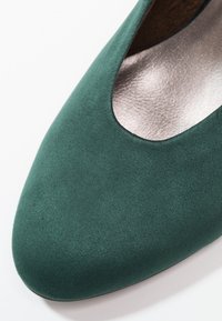 Grön mockasinsko med rund tå och utklippt detalj upptill. Slät silvrig insida med platt sula. Inga synliga beslag eller accenter.