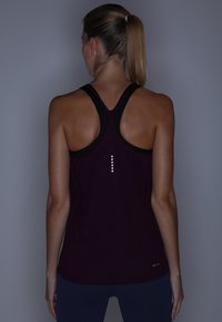Femme avec une queue de cheval blonde portant un débardeur de sport noir dos nageur et un legging foncé, debout, dos à la caméra.