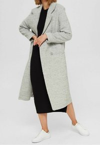 Woman wearing a gray textured long coat over a black ribbed midi dress and white sneakers, standing with hands near waist.