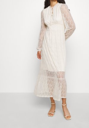 Kvinde iført en hvid blonde midi-kjole med lange, gennemsigtige ærmer og åben tå sandaletter med ankelrem, stående mod en ensfarvet hvid baggrund.