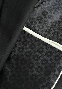 Schwarzer Jackenstoff mit einem strukturierten Muster. Das Innenfutter ist glatt und mit einem geometrischen Design versehen, akzentuiert durch einen weißen Rand.