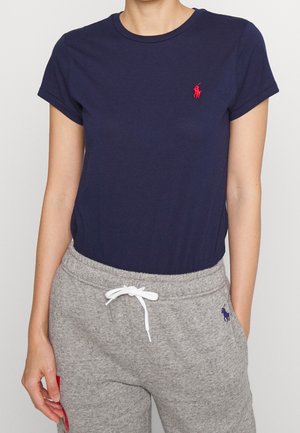 Kvinde iført en navyblå tætsiddende T-shirt med rødt logo og grå joggingbukser med hvid snor og navyblåt logo, stående med den ene hånd i lommen.