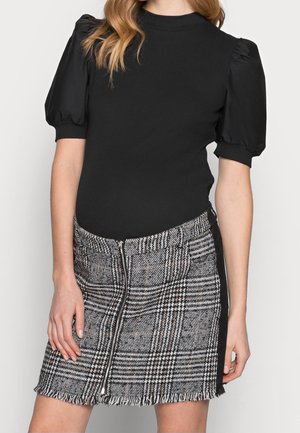 Mujer que lleva un top negro con mangas abullonadas y una minifalda de cuadros en blanco y negro con cremallera frontal y borde con flecos.