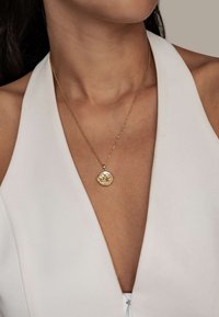 Gold pendant necklace with a circular charm featuring intricate details, hanging on a fine chain, worn over a white garment with a deep neckline.