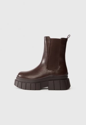 Bottines en cuir marron foncé avec une semelle épaisse et chunky et un panneau latéral en élastique, sur fond blanc uni.