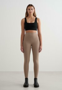 Melna cropped blūze un bēšas augstas vidukļa legingu bikses, kas izgatavotas no elastīga materiāla, apvienotas ar melniem potīšu zābakiem, uz neitrāla fona.