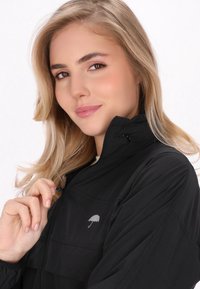 Svart zip-up jacka med en hög krage och en subtil paraplylogotyp. Tyget verkar lätt och har en mjuk textur.