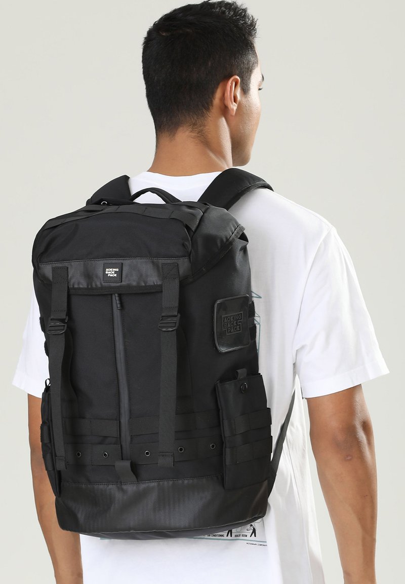 Schwarzer Stoff-Rucksack mit verstellbaren Trägern, Seitentaschen und einem strukturierten unteren Bereich. Enthält ein Logo-Patch auf der Oberseite.