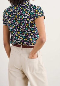 Femme portant une chemise à manches courtes à fleurs violettes, jaunes et blanches, rentrée dans un pantalon beige avec une ceinture brune tressée.