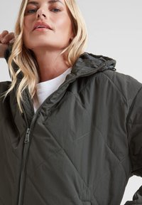 Blonde woman wearing a dark green quilted jacket with hood, partially zipped, and a white shirt underneath against a plain background.