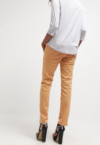 Pantaloni beige chiari a vita alta con vestibilità slim, dotati di tasche posteriori e una texture liscia, abbinati a tacchi alti neri.