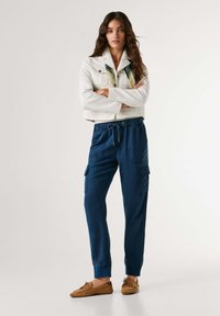 Žena s dlhými vlasmi stojí so založenými rukami, oblečená v bielej skrátenej bunde, modrých cargo teplákoch a hnedých mokasínových mokasínach.